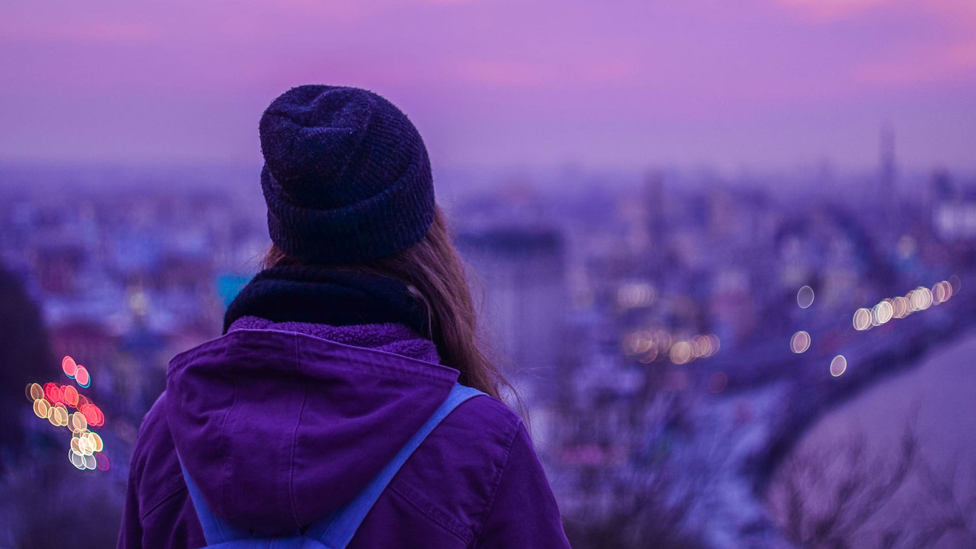 Kuvassa on violettiin takkiin pukeutunut nainen, nähtynä takaapäin, katsomassa kaupunkiympäristöä.
